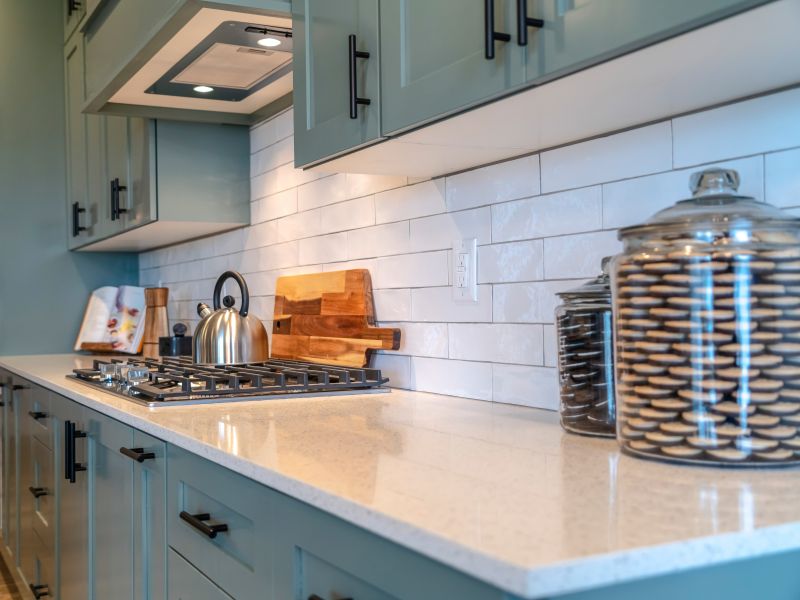 Kettle on cooktop fixed on white countertop with cookie jars inside home kitchen. Exhaust hoos, hanging cabinets, and cooking appliances can also be seen inside the room of a california, md kitchen.