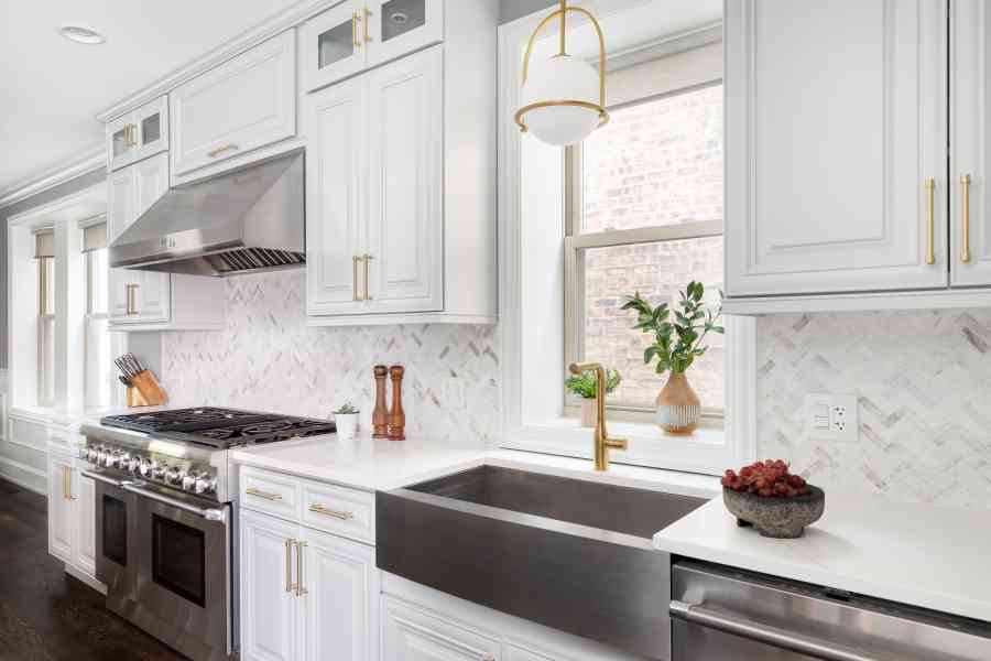 A kitchen with a gold light fixture hanging over a stainless steel apron sink and gold faucet, white marble countertop, and a white marble herringbone tile backsplash of a leonardtown maryland home.
