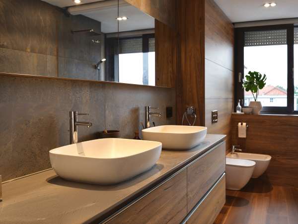 A stylish modern bathroom featuring natural wood elements, sleek white basins, and contemporary fixtures. The design includes a gray stone wall, wooden floors, and a large window providing natural light.