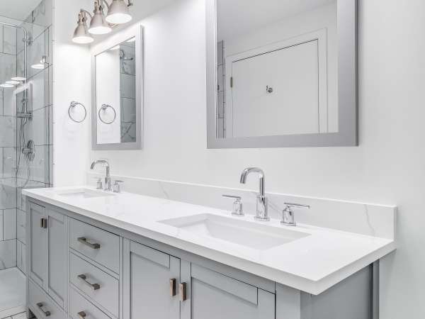 An elegant, remodeled bathroom with a grey vanity and bronze hardware. The shower has a large shower head and marble tiles and glass wall line the sides.